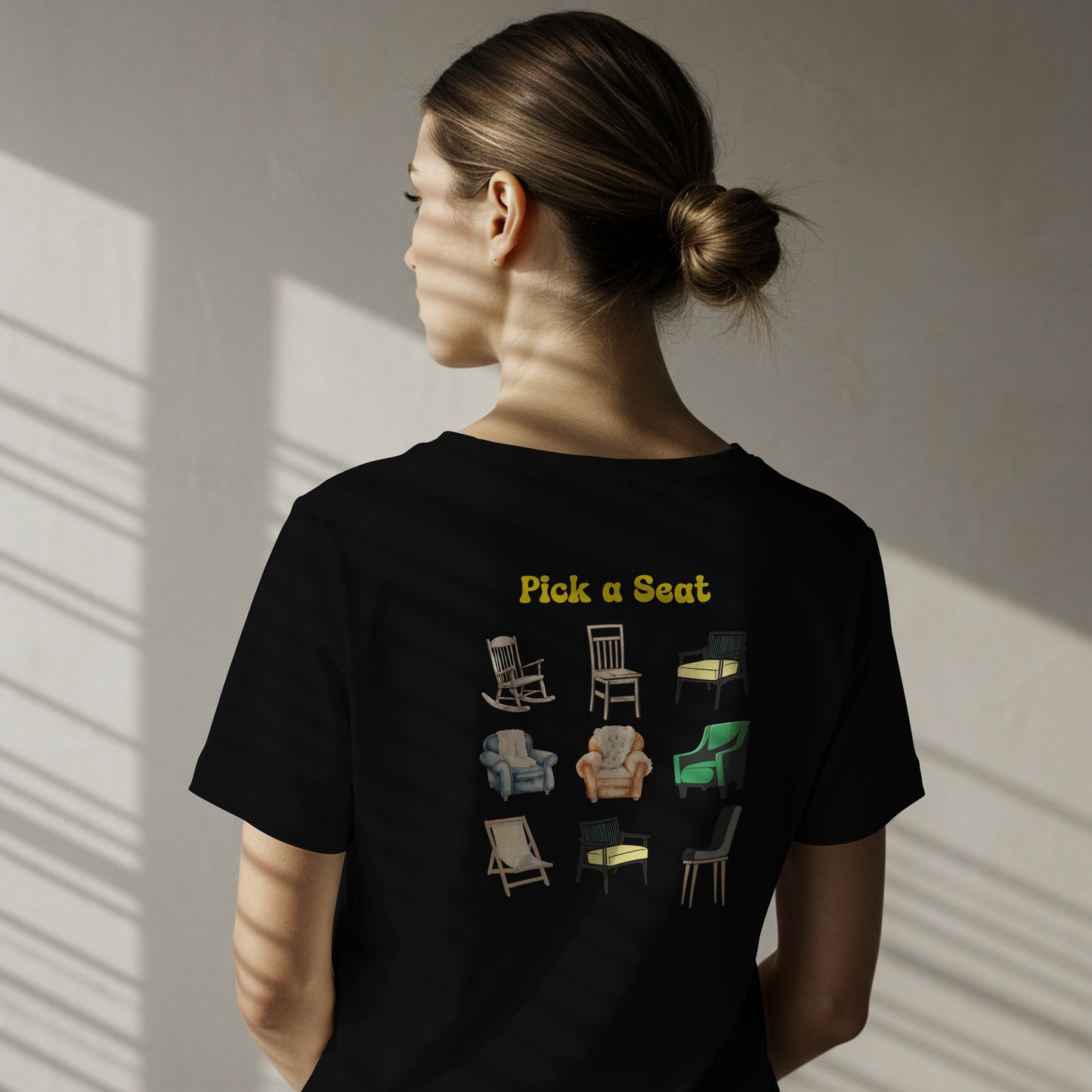 Person wearing a black t-shirt with Illustrations of various chairs with the text 'Pick a Seat' above them on a white background. The graphic is on the back.
