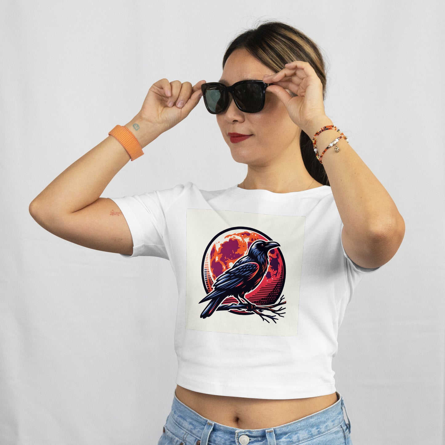 Woman wearing a white t-shirt with a black crow perched on a branch with an orange and red moon in the background. The model is holding sunglasses.