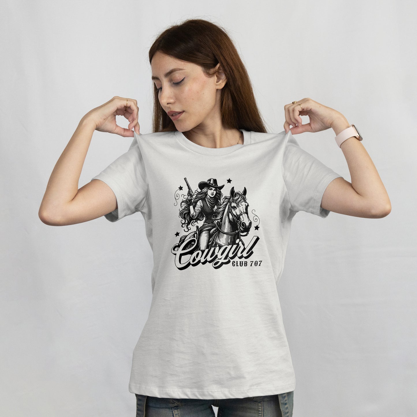 Woman wearing a white t-shirt with a black and white illustration of a cowgirl on a horse with 'Cowgirl Club 707' text. The model is standing on a plain background