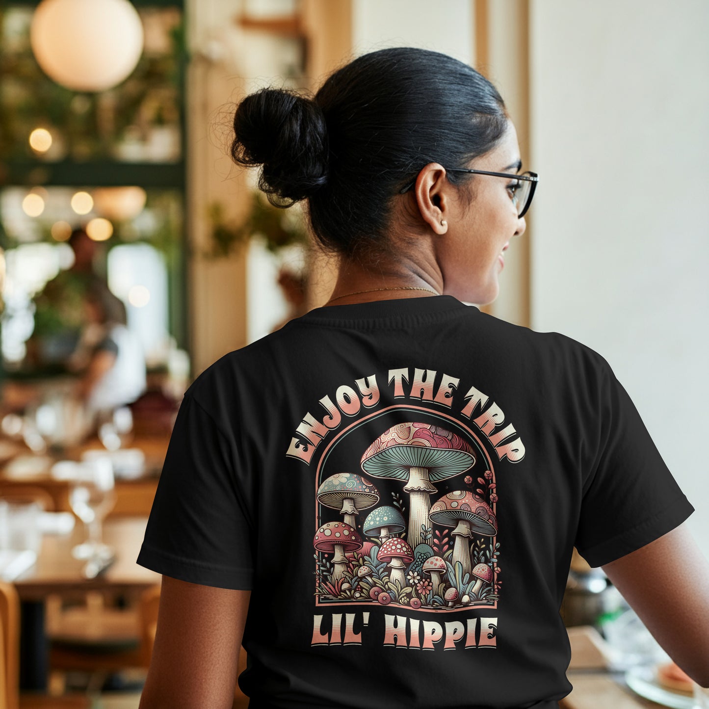 Person wearing a black t-shirt with a colorful illustration of mushrooms with 'Enjoy the Trip, Live Hippie' text on a white background, in a restaurant setting