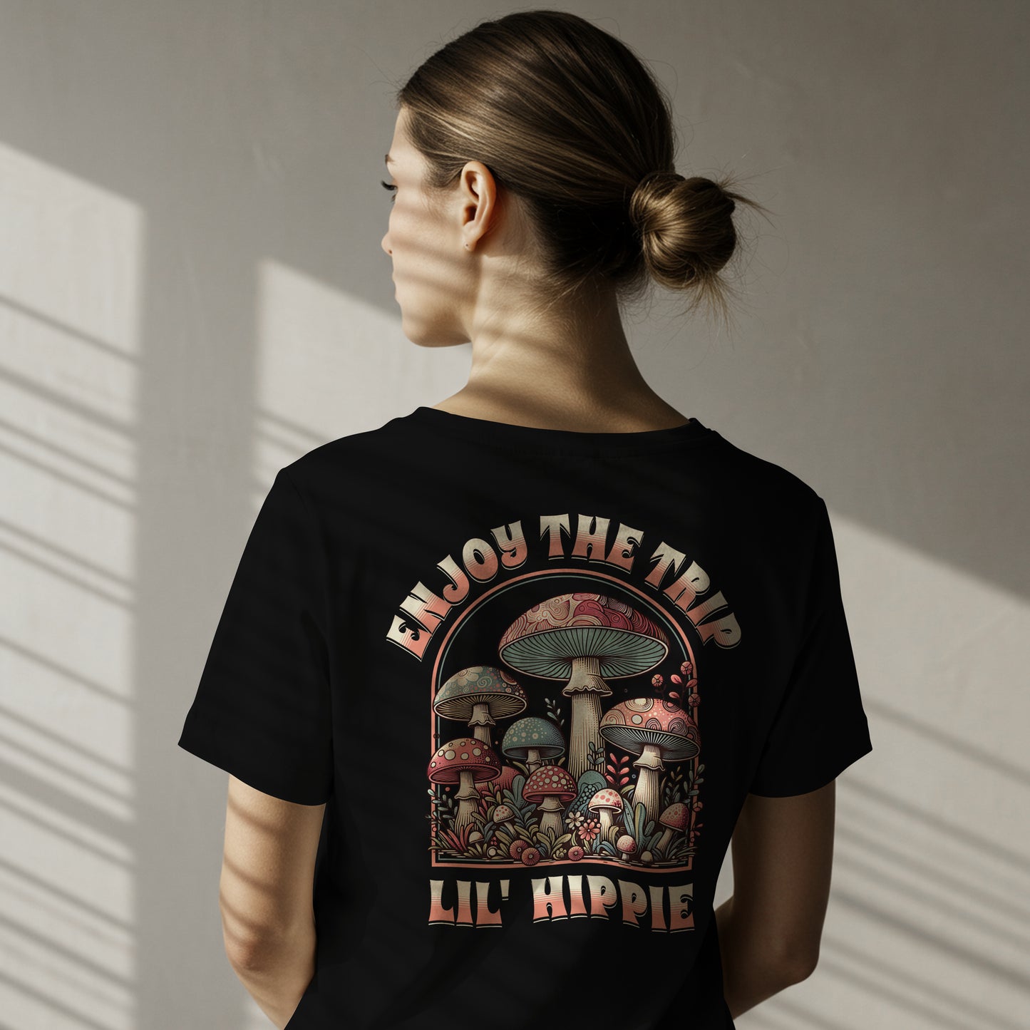 Person wearing a black t-shirt with a colorful illustration of mushrooms with 'Enjoy the Trip, Live Hippie' text on a white background. The graphic is on the back of the shirt.
