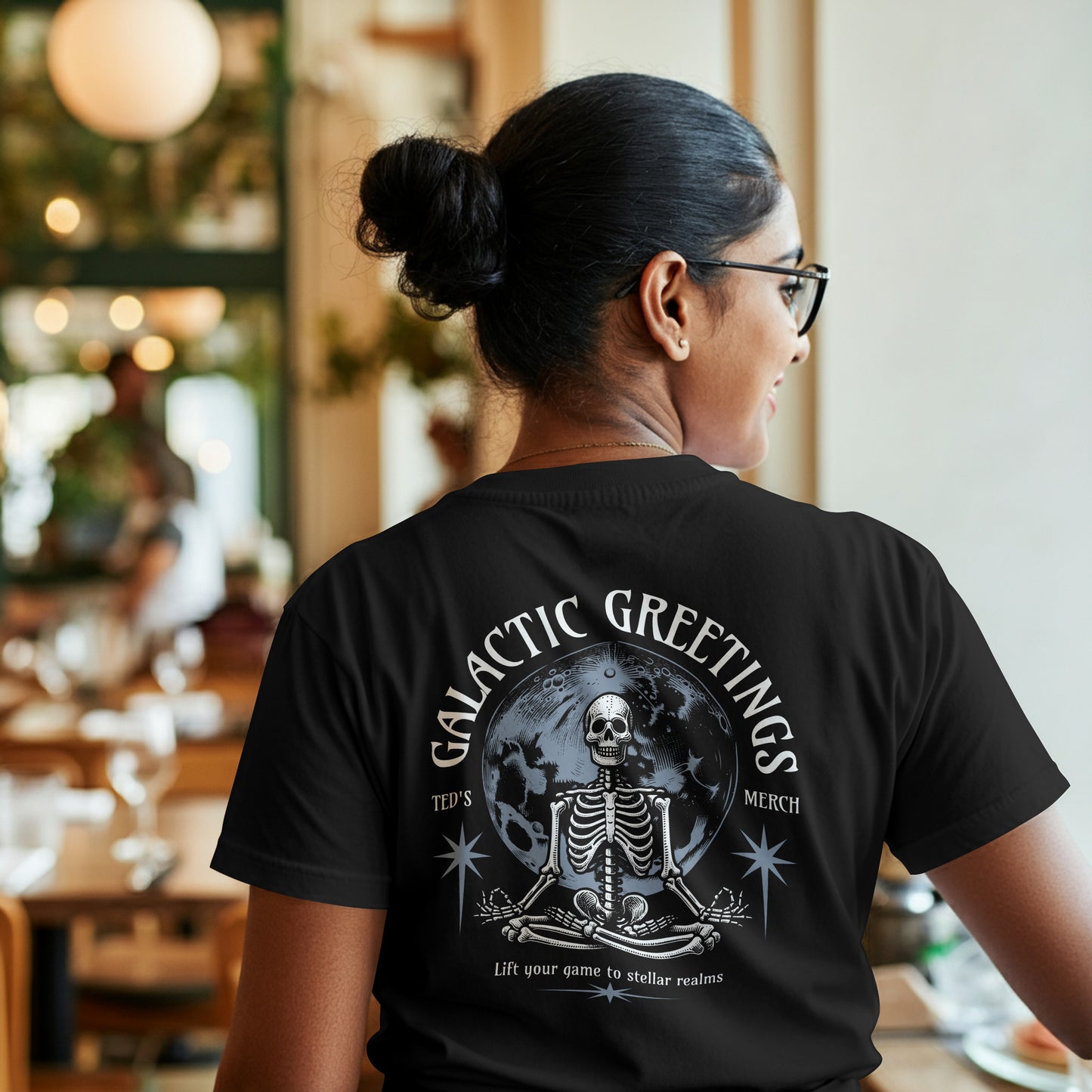 Person wearing a black t-shirt with skeletal design with 'Galactic Greetings' text and moon in center on black background. The model is in a restaurant setting.