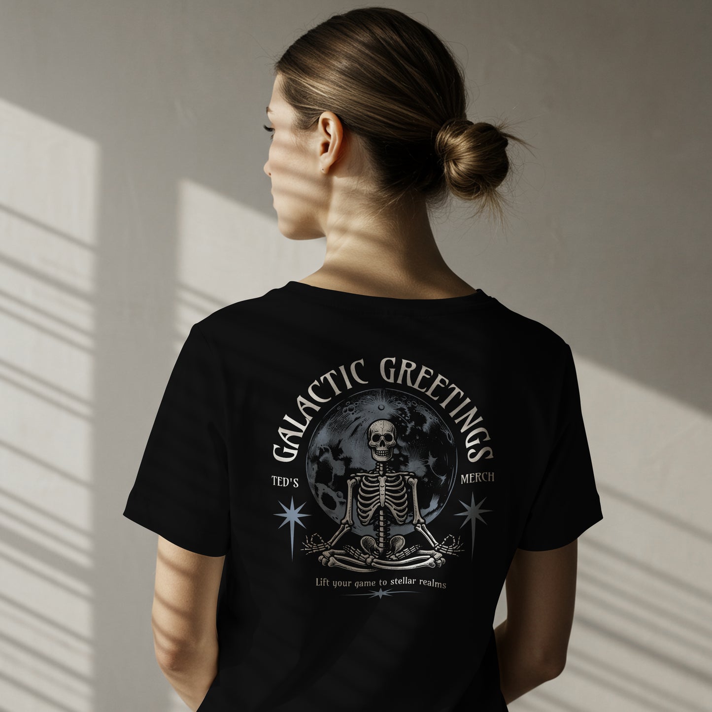 Person wearing a black t-shirt with skeletal design with 'Galactic Greetings' text and moon in center on black background on the back. The model standing against a neutral wall.