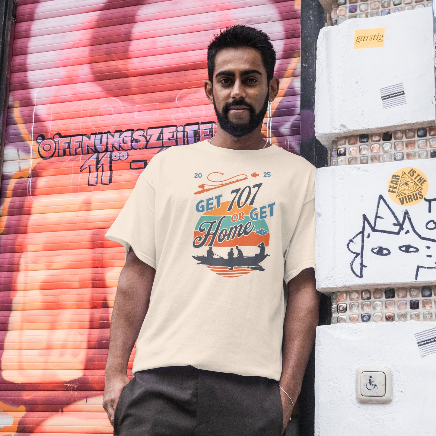 Man wearing a t-shirt with a motivational graphic with 'Get 707 or Get Home' text and silhouette of people in a boat. in front of a colorful mural.