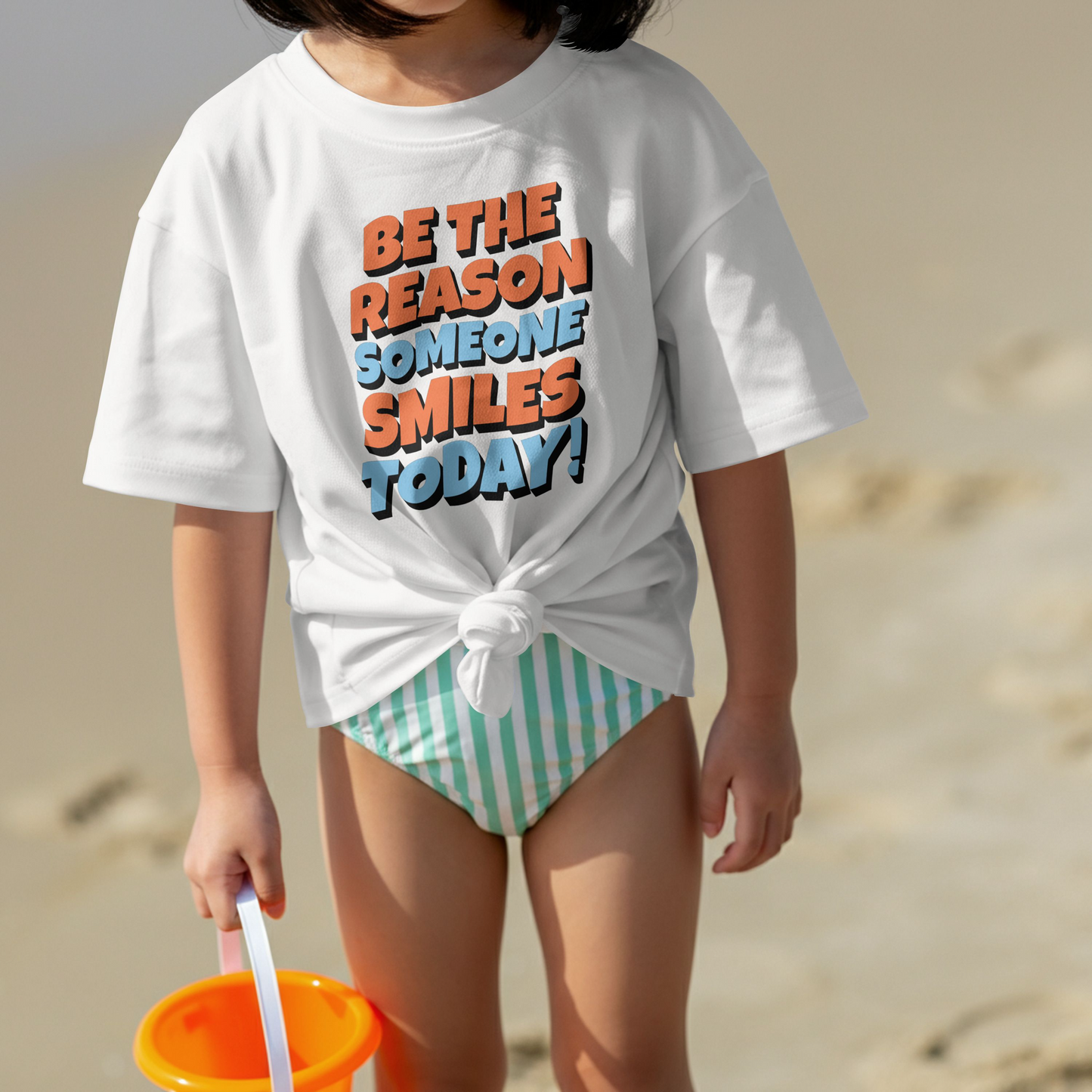 Child wearing a white t-shirt with Motivational quote 'Be the reason someone smiles today!' in orange and blue on a white background. The child has striped shorts on and is on a beach.