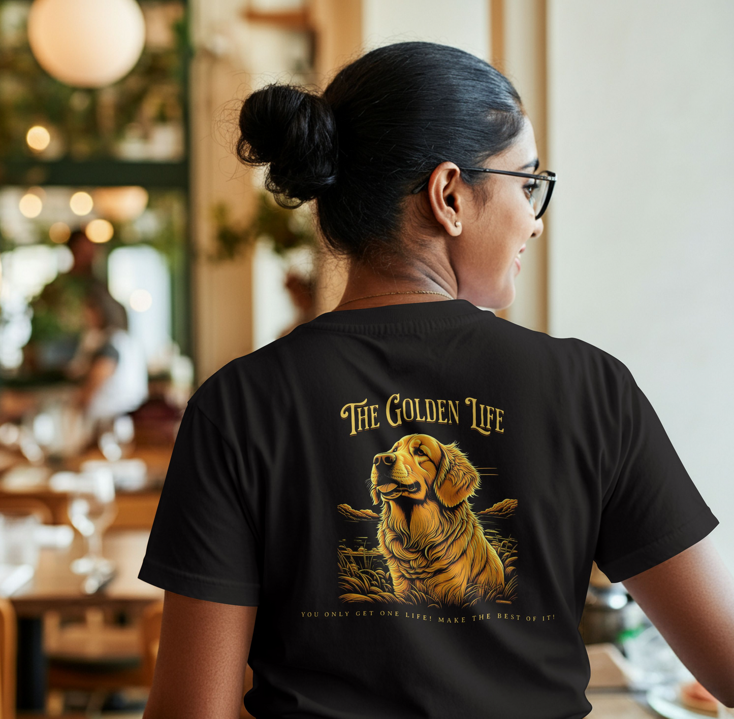 Person wearing a black t-shirt, with a illustration of a golden retriever with 'The Golden Life' text on a black background. The person is in a restaurant setting.