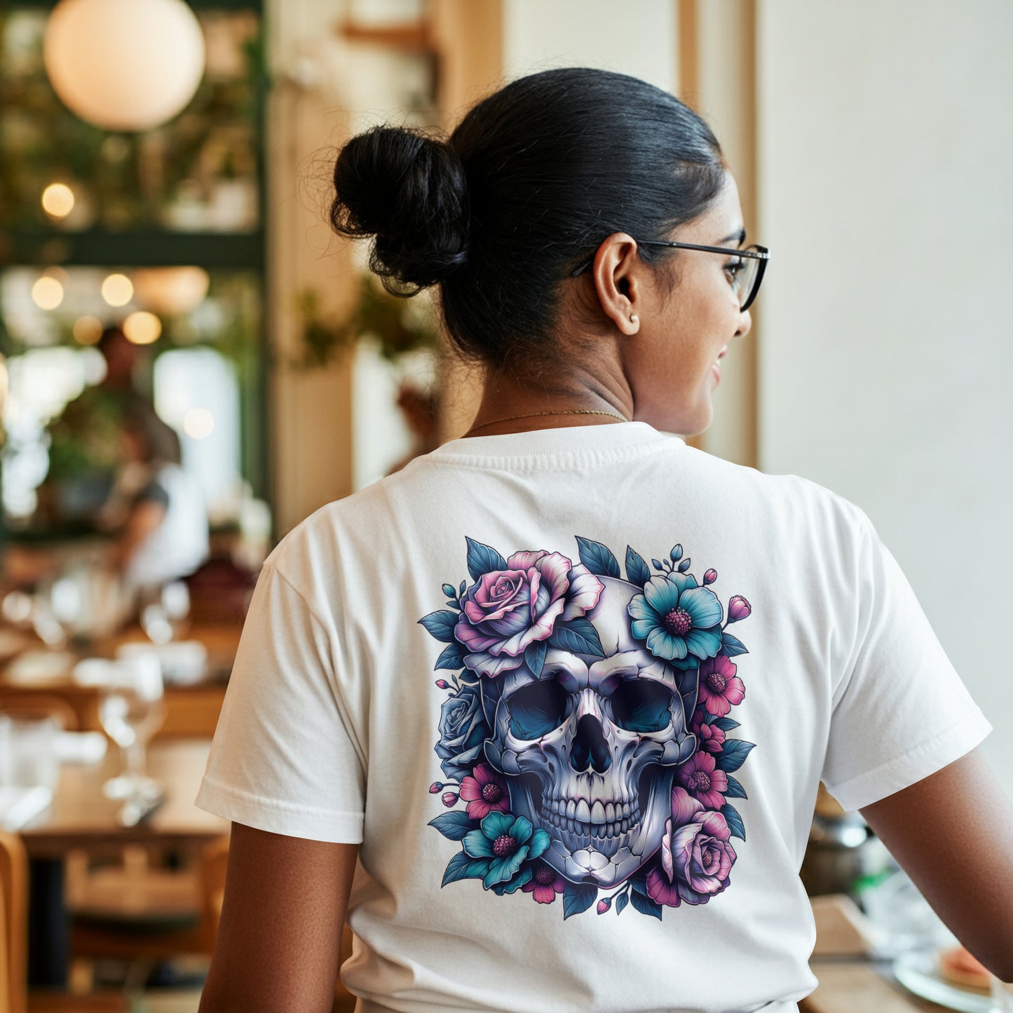 Person wearing a white t-shirt with a Colorful skull design with pink and blue flowers on a white background on the back of the shirt. The model is standing in a restaurant.