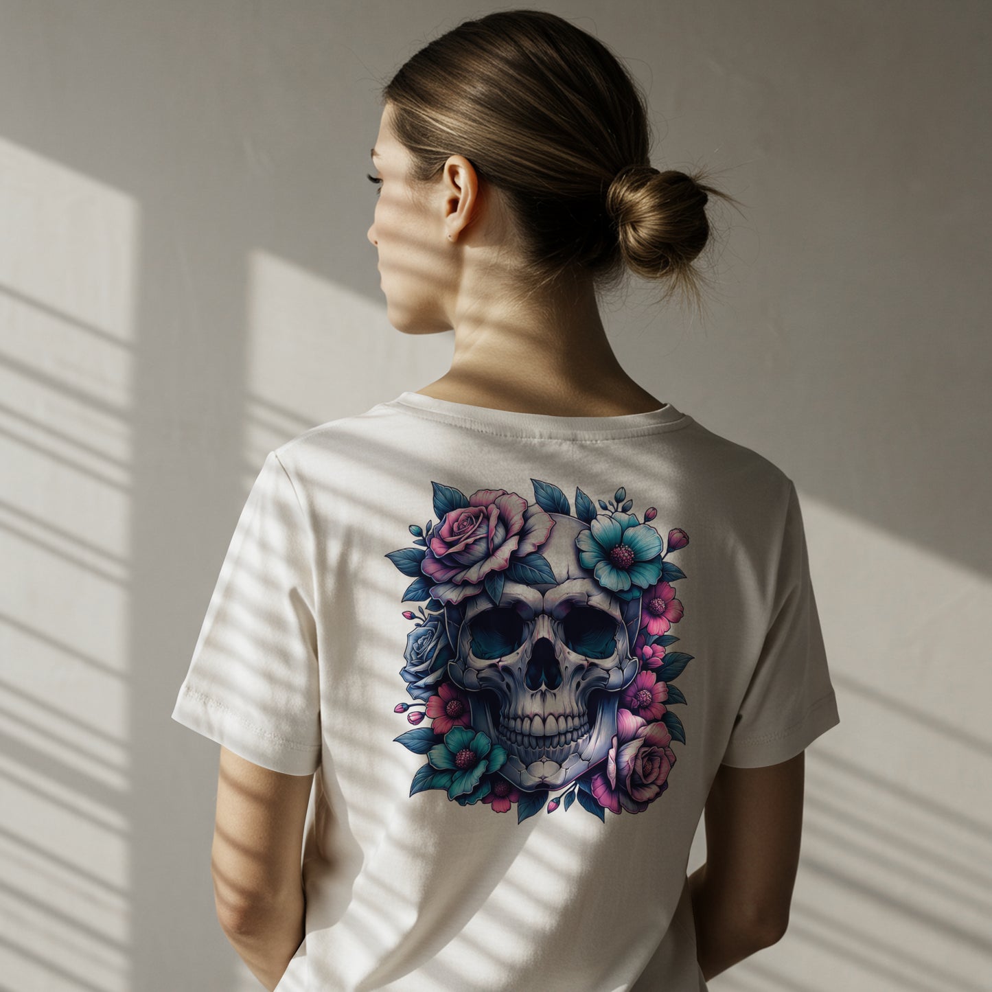 Person wearing a white t-shirt with a colorful skull design with pink and blue flowers on a white background on the back of her shirt. The model is standing against a neutral wall.
