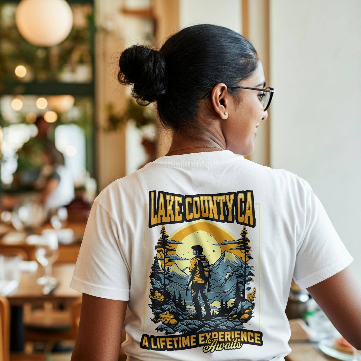 A woman wearing a white graphic T-shirt with a design featuring a person looking at a mountain range with the text 'LAKE COUNTY CA' and 'A LIFE TIME EXPERIENCE AWAITS'. The background is in shades of blue and yellow, creating a vibrant contrast. The model is sitting in a restaurant setting.