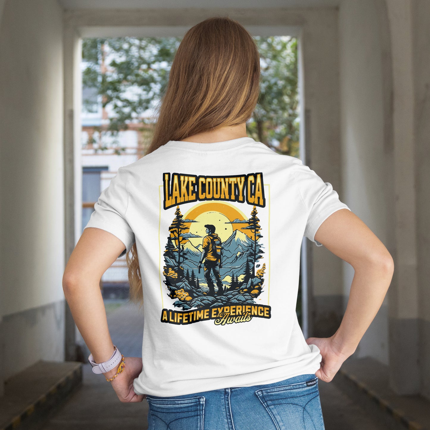 Person wearing a graphic T-shirt with a design featuring a person looking at a mountain range with the text 'LAKE COUNTY CA' and 'A LIFE TIME EXPERIENCE AWAITS'. The background is in shades of blue and yellow, creating a vibrant contrast. The graphic is on the back. The model is standing in a room with a window in the background.