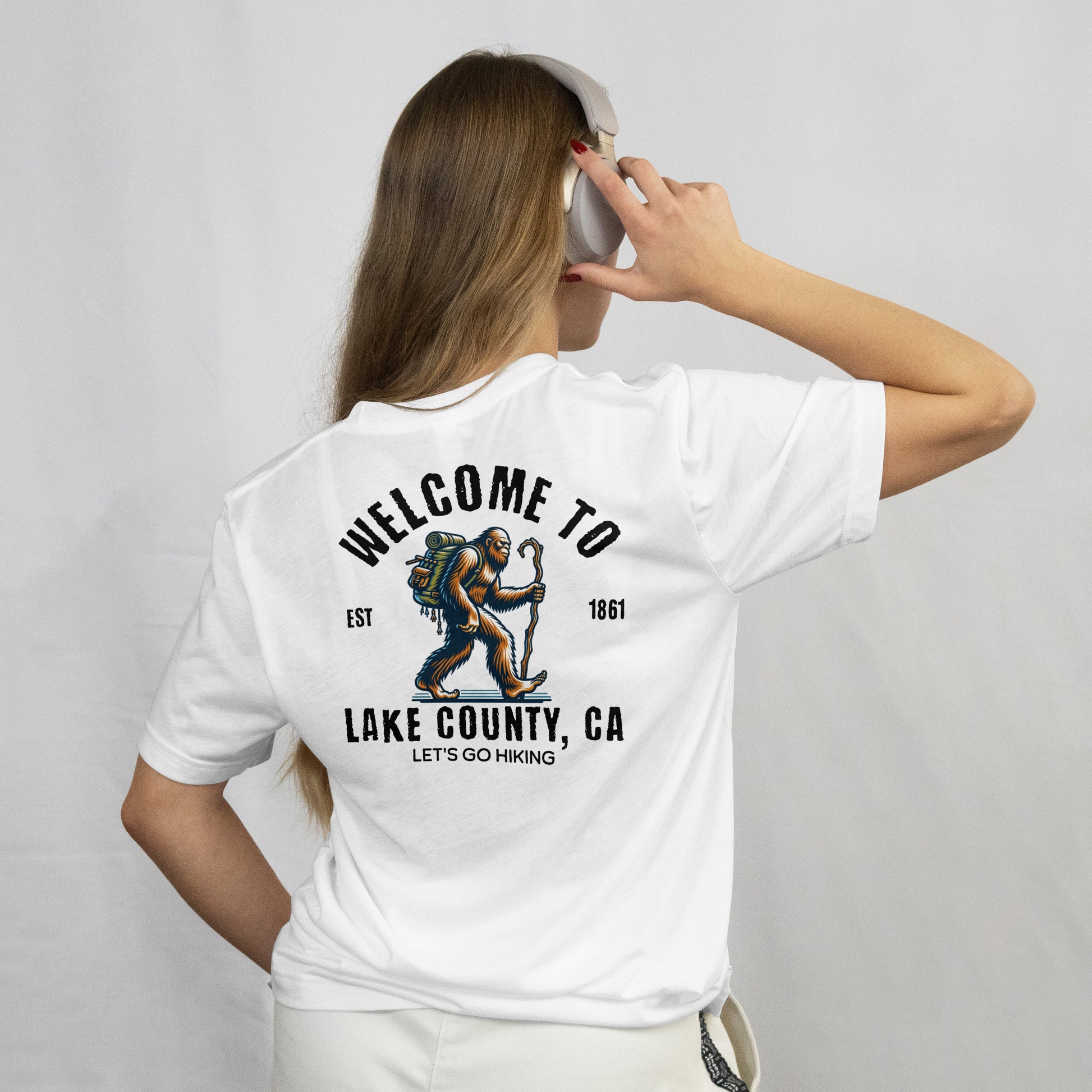 Person wearing a white t-shirt with A graphic image of a hiking Sasquatch character with a backpack and a staff. The text welcome to appear at the top and Lake County CA at the bottom. In smaller font saying lets go hiking on the back.