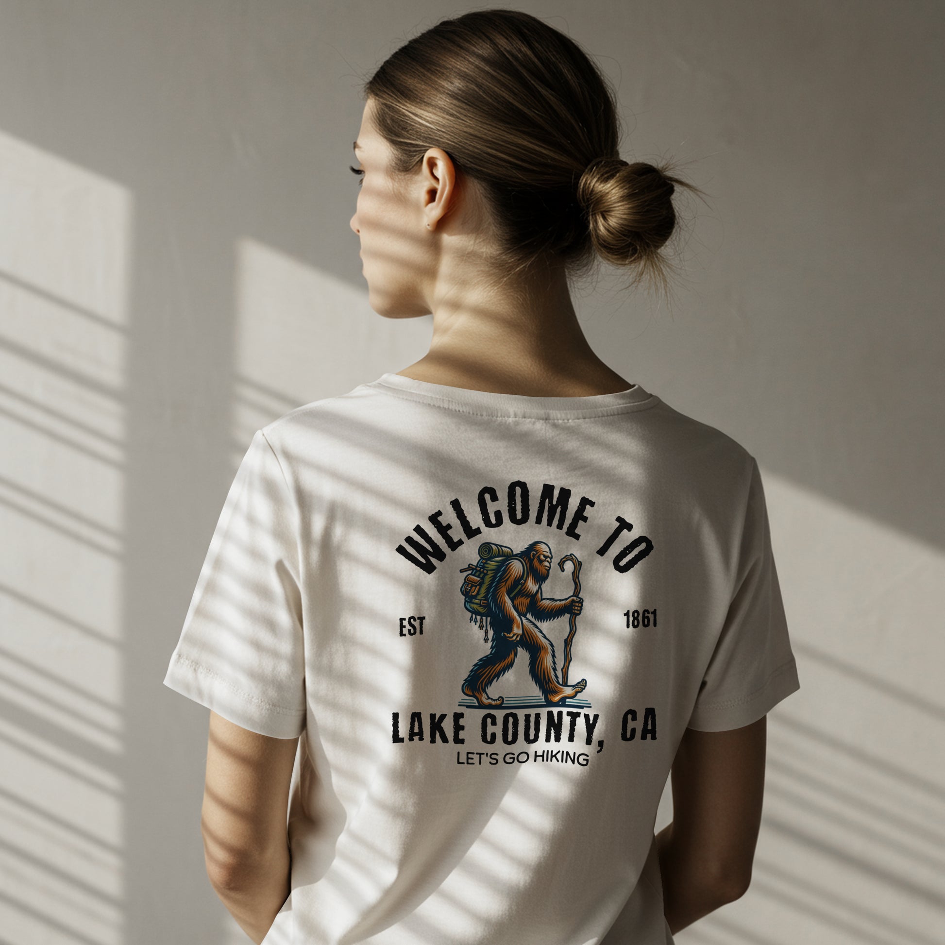 Person wearing a white t-shirt with a A graphic image of a hiking Sasquatch character with a backpack and a staff. The text welcome to appear at the top and Lake County CA at the bottom. In smaller font saying let's go hiking on the back, standing against a neutral wall.