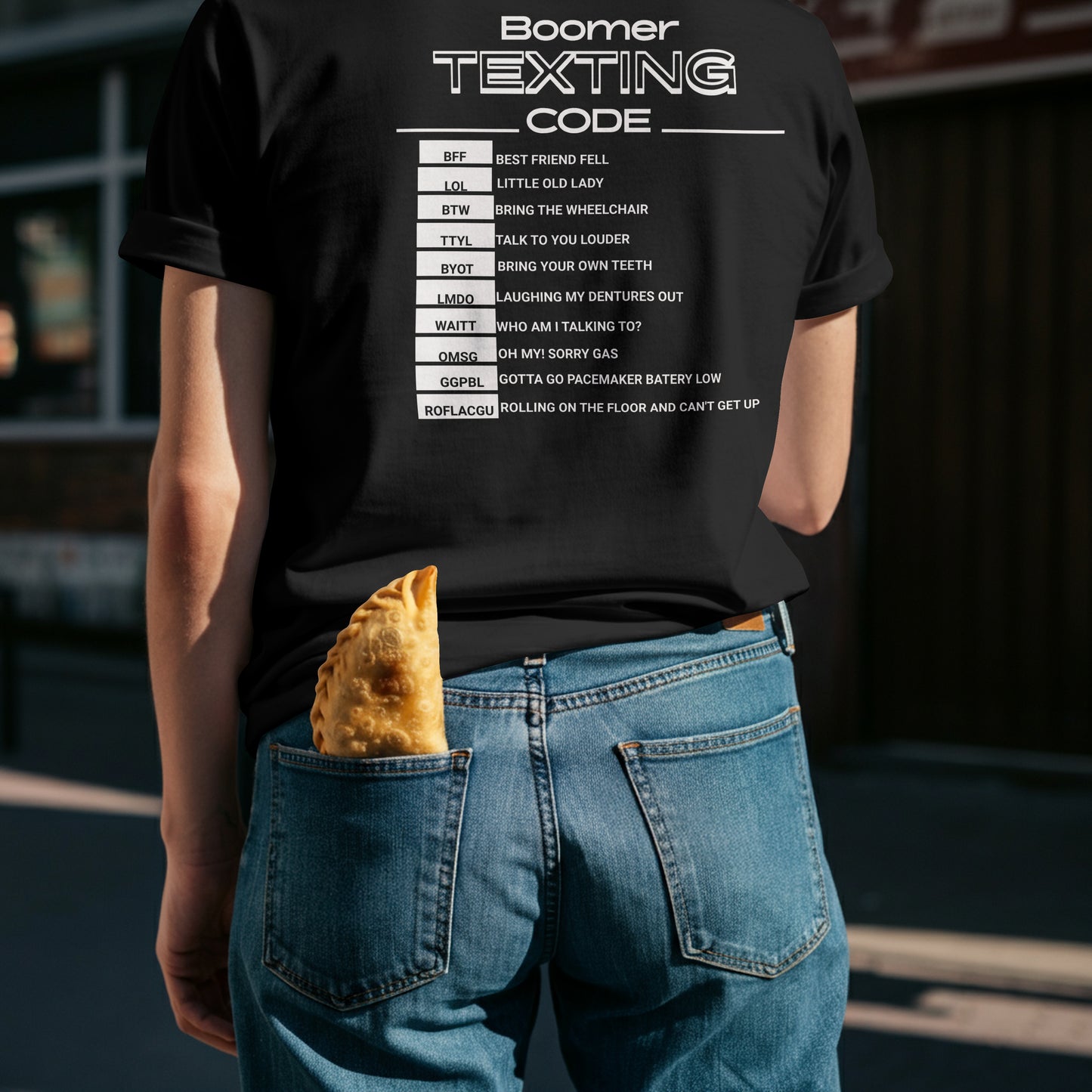Person wearing a black t-shirt with 'Boomer Texting Code, The Code consists of abbreviations and there means. the model is standing outdoors.