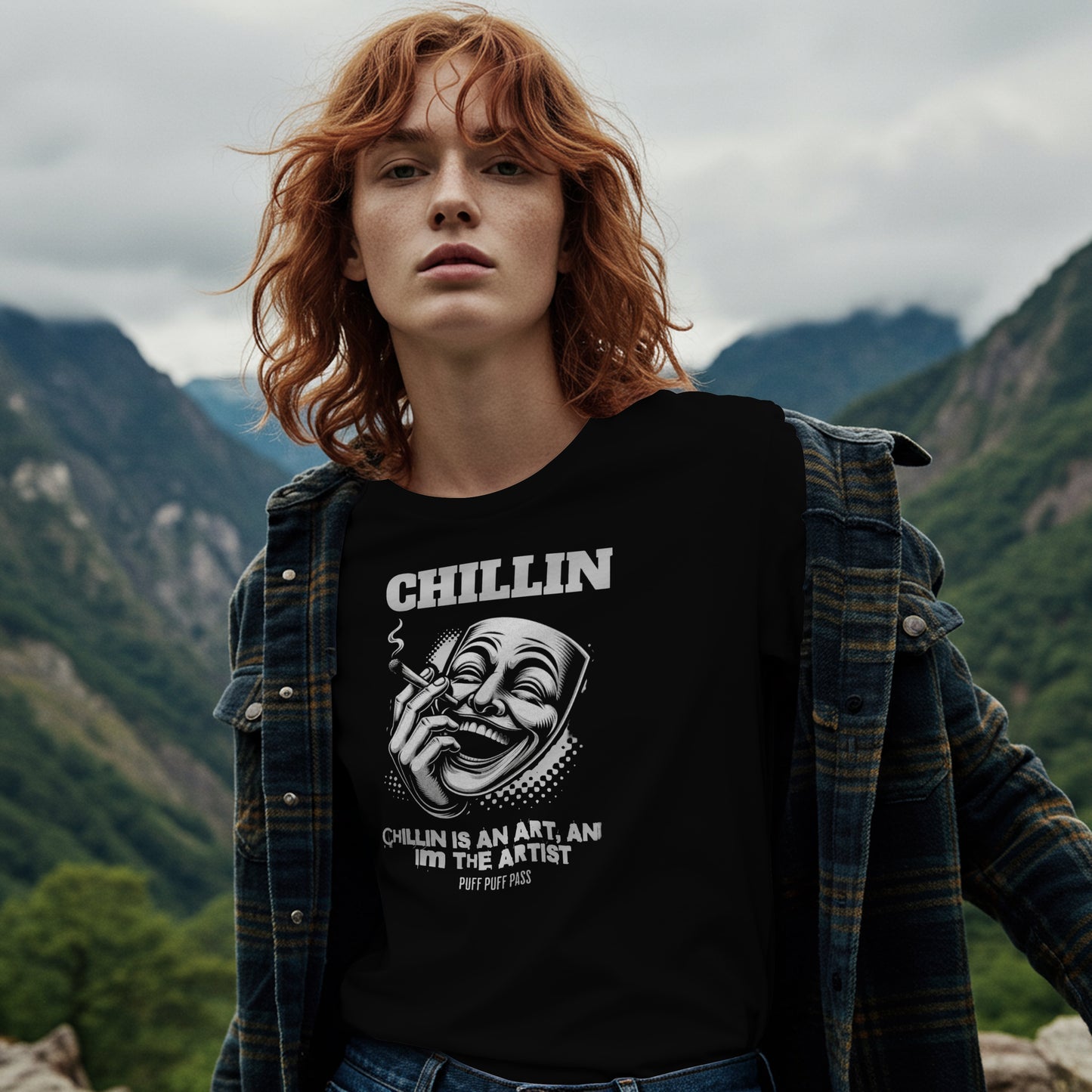 Person wearing a black t-shirt with Black and white illustration of a mask with text 'Chillin is an art, I'm the artist' on a black background. The model is standing in a mountainous landscape
