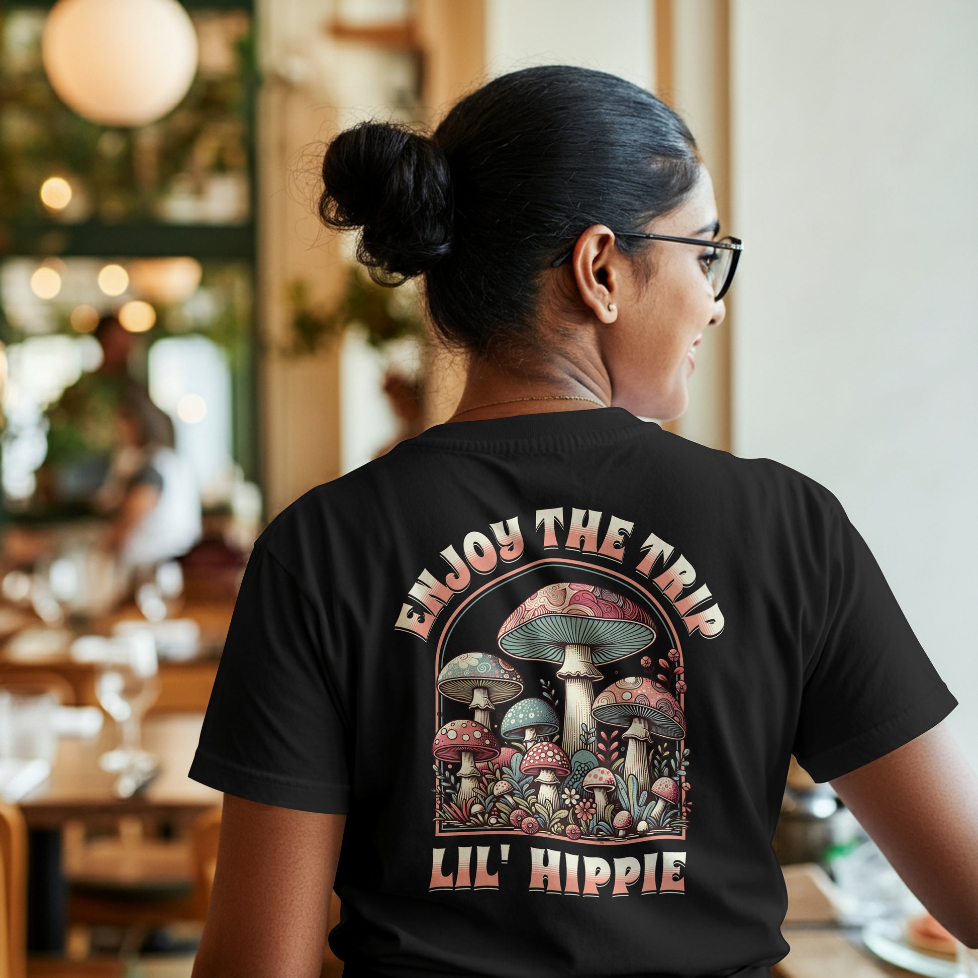 Person wearing a black t-shirt with a colorful illustration of mushrooms with 'Enjoy the Trip, Live Hippie' text on a white background, in a restaurant setting