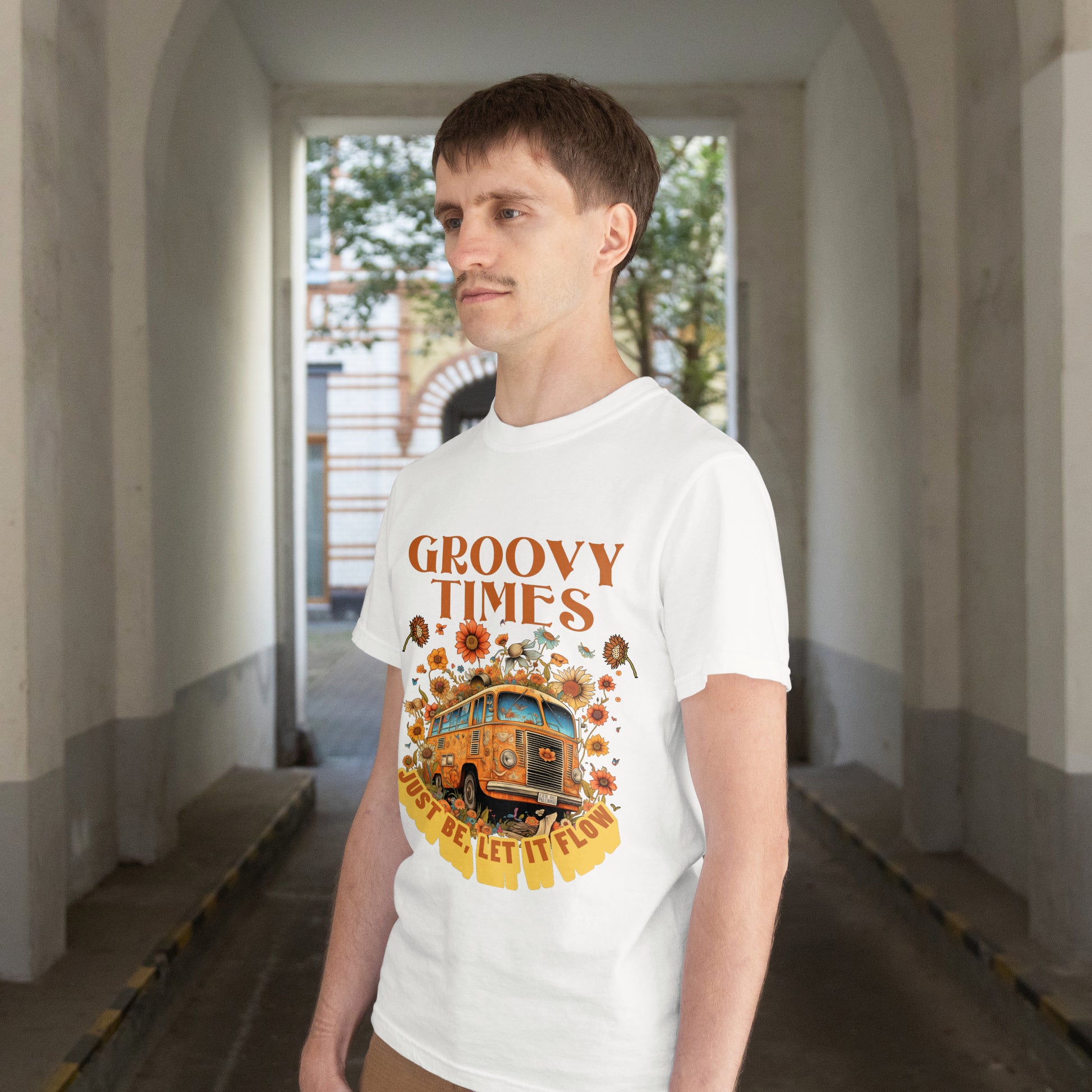 Person wearing a white t-shirt with a Vintage bus surrounded by flowers with 'Groovy Times Just Be, Let It Flow' text on a white background in an indoor setting.