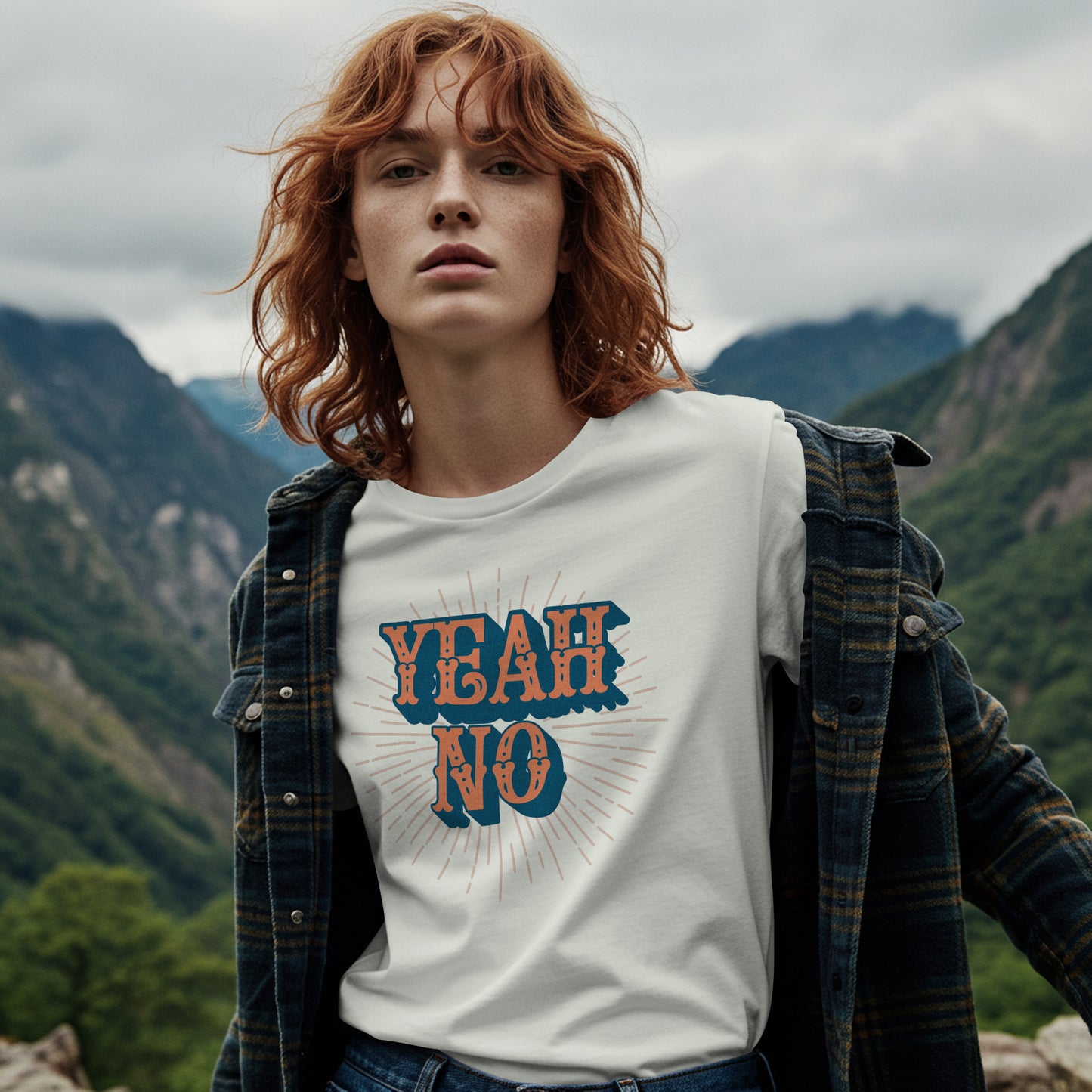 Person wearing a white t-shirt with Text 'YEAH NO' in blue and orange on a white background with sunburst design, in a mountainous landscape